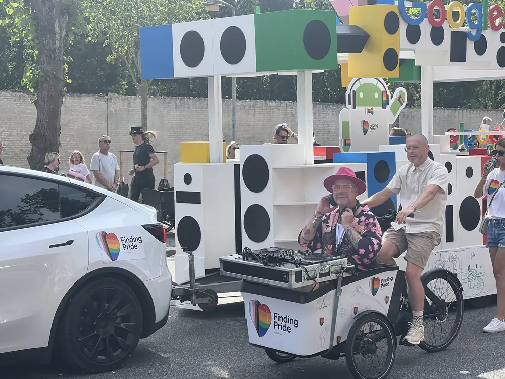 Ladcykel med DJ-pult og højttalere på ladet, med paradevogn i baggrunden.