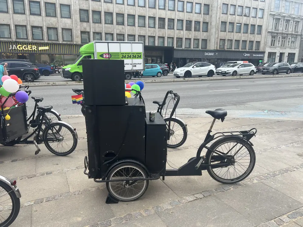 Sort ladcykel med lydanlæg og batterikasse monteret på ladet, parkeret på en gade i København.
