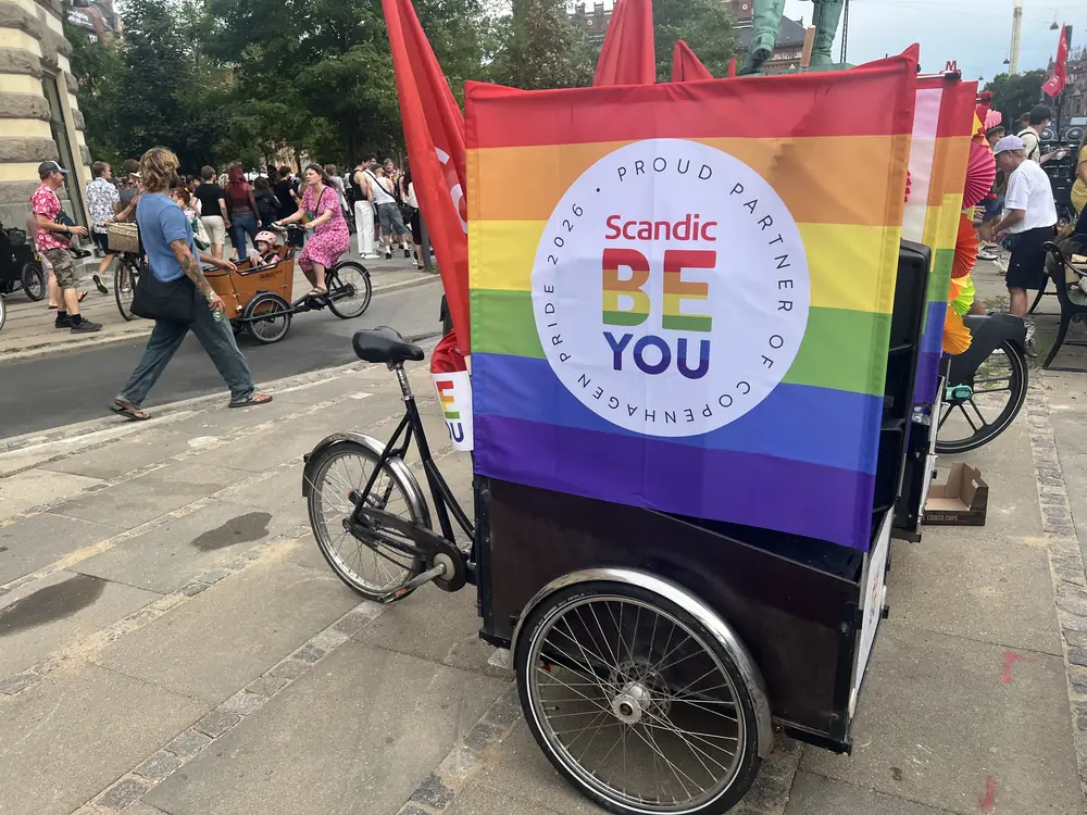 Ladcykel med regnbuefarvet banner og flag, holdende ved ruten før afgang.